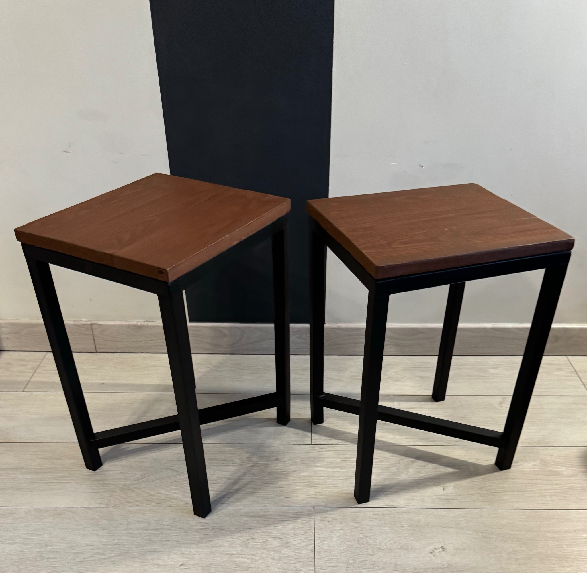 Dwa identyczne stołki barowe z kwadratowymi siedziskami z ciemnego drewna i czarnymi metalowymi nogami o geometrycznym kształcie na tle ściany w biało-czarne pasy.