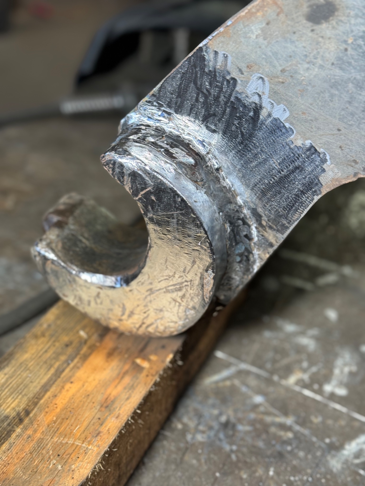 Fragment metalowego elementu z widocznymi śladami spawania, leżącego na drewnianym podłożu, ukazujący proces łączenia metali.