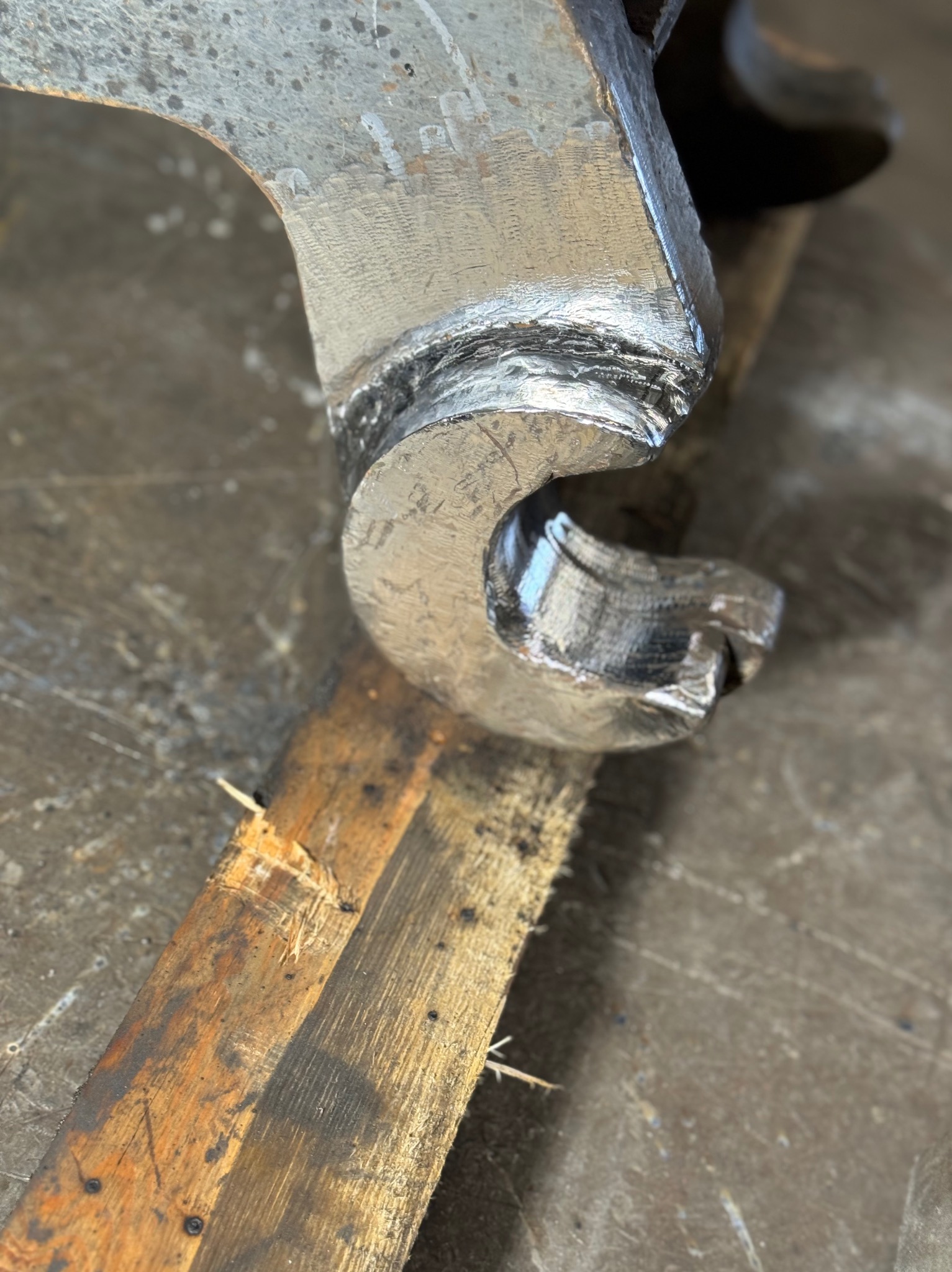 Szlifowany element metalowy o zakrzywionym kształcie, leżący na desce z widocznymi śladami obróbki.