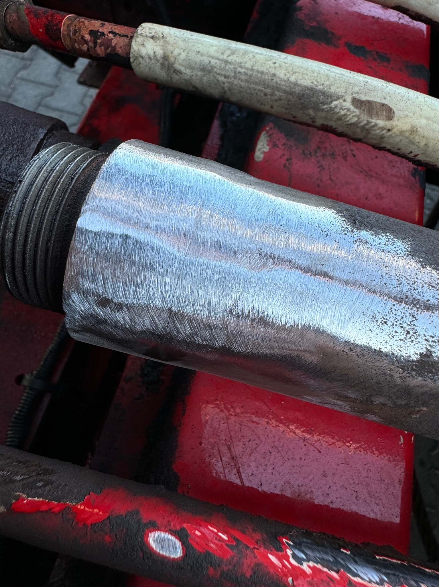 Szlifowany, gwintowany metalowy element konstrukcyjny, odbijający światło, umieszczony na czerwonej, pomalowanej konstrukcji.