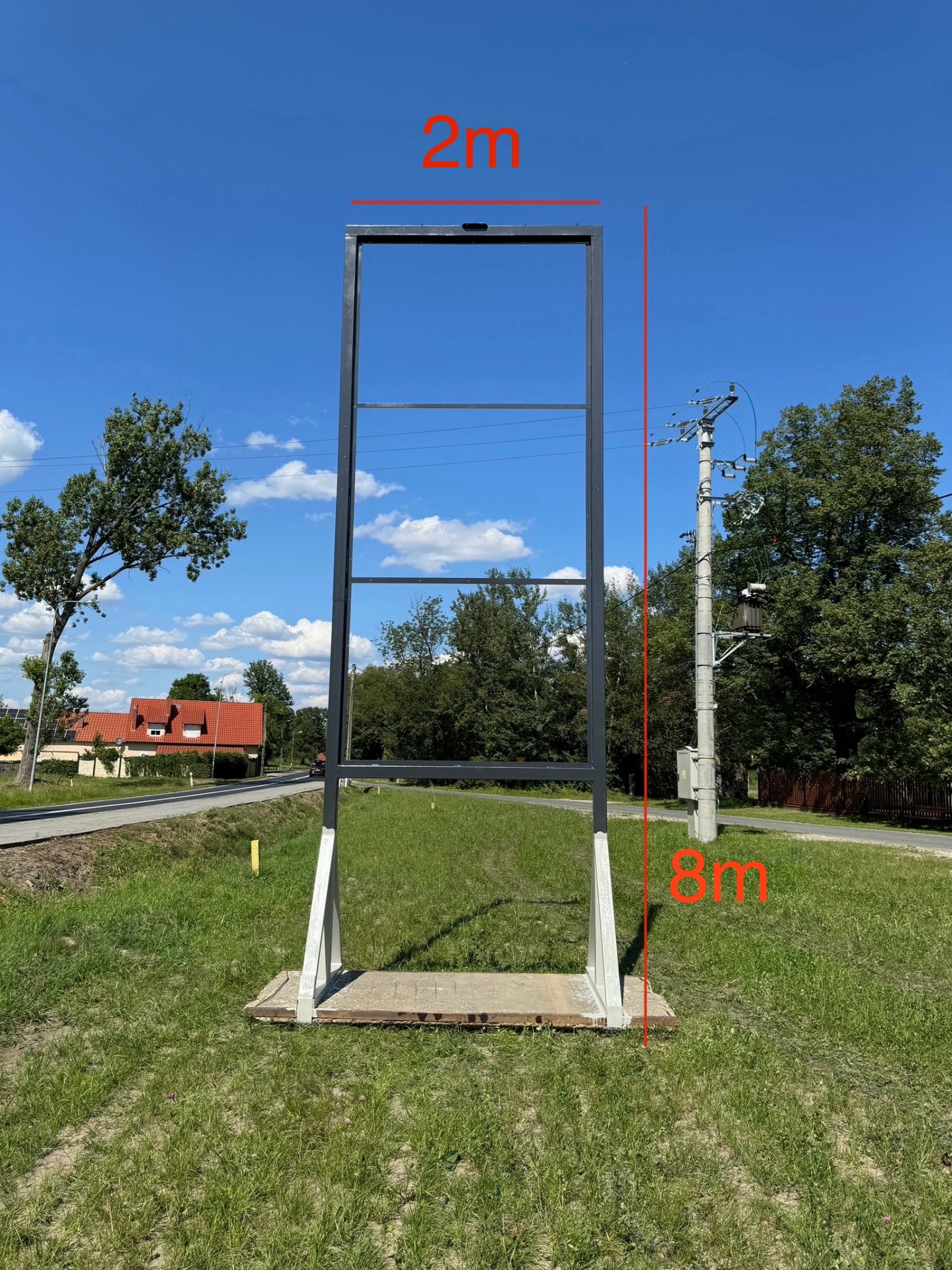 Wysoka, metalowa konstrukcja ramy, prawdopodobnie pod billboard lub reklamę, z widocznymi wymiarami 2m x 8m, umieszczona na betonowej podstawie na trawiastym terenie, z budynkami i słupem...