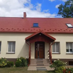 Wykończony dom jednorodzinny z czerwoną dachówką, kremową elewacją, oknami dachowymi i zadaszonym wejściem z drewnianymi elementami, otoczony zielenią w słoneczny dzień.