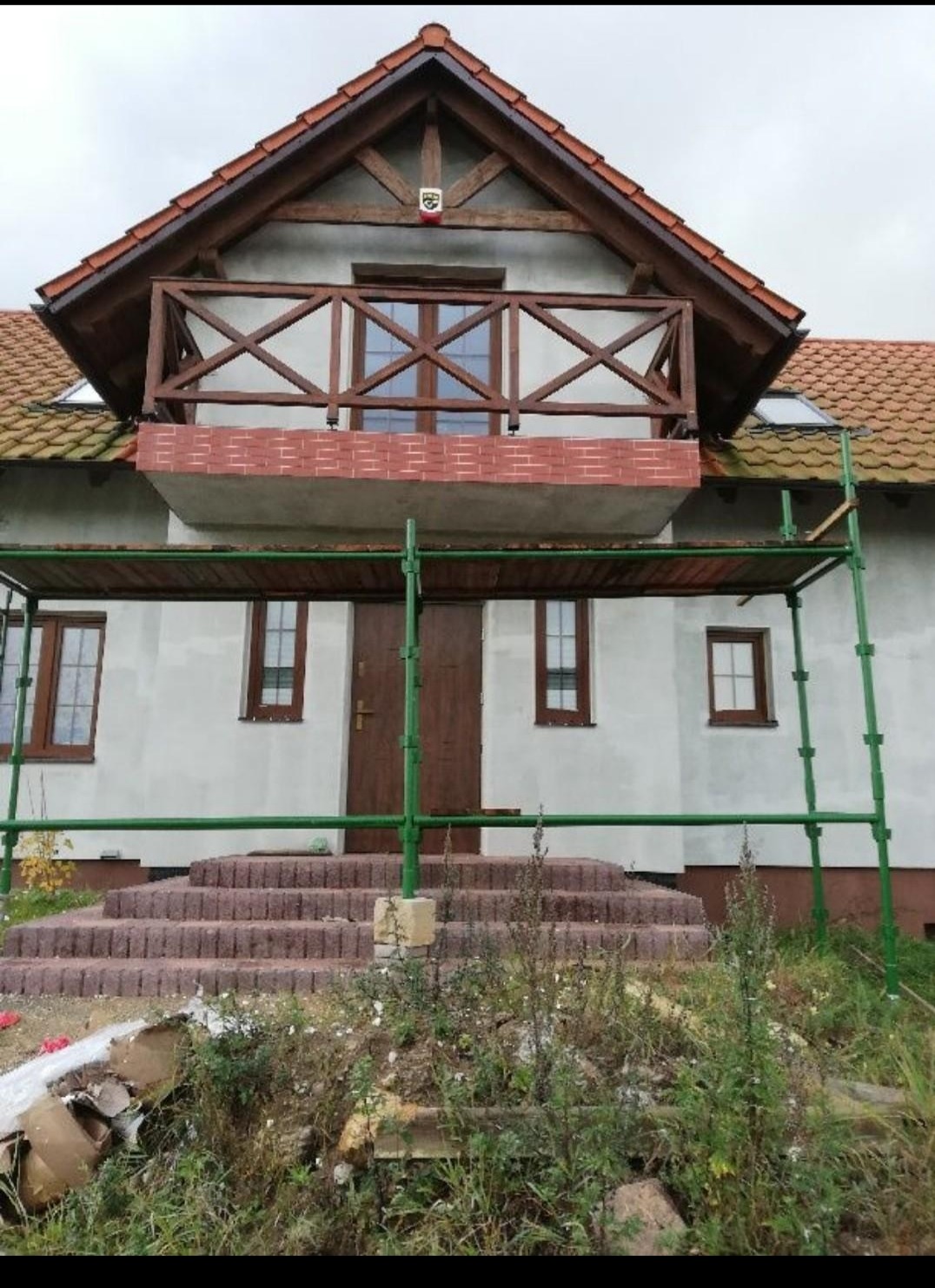 Dom z balkonem w trakcie budowy, widoczne rusztowanie, schody wejściowe z kostki, niezagospodarowany teren przed domem, herb na szczycie dachu.
