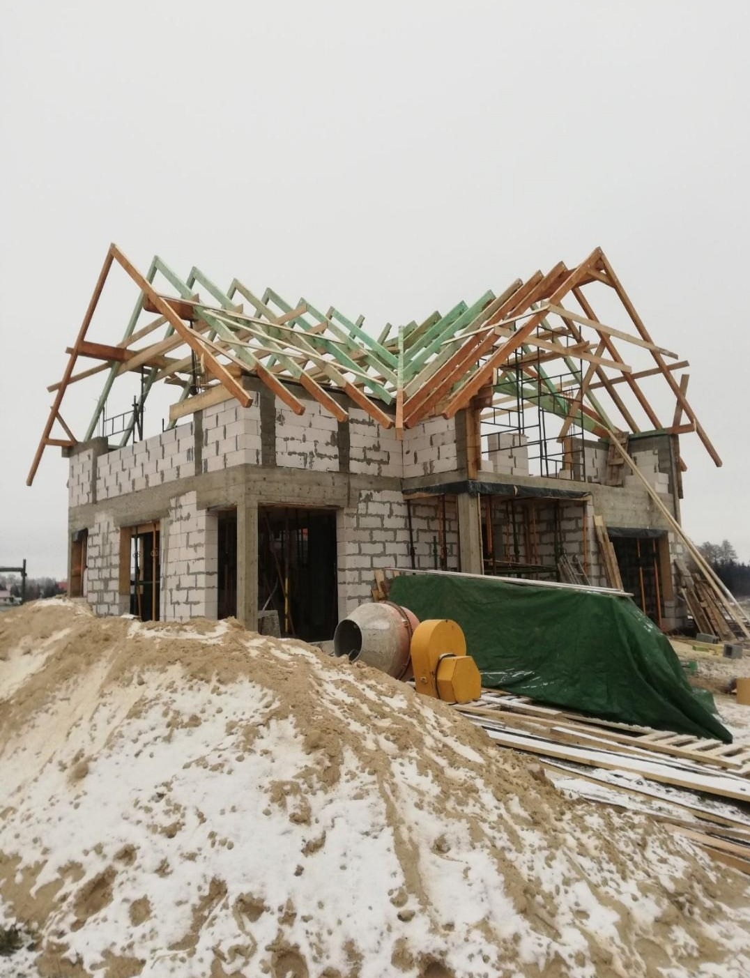 Surowy stan domu z białego bloczka, z częściowo ukończoną drewnianą konstrukcją dachu, widok z poziomu ziemi z hałdą piasku i sprzętem budowlanym na pierwszym planie, w zimowej scenerii.