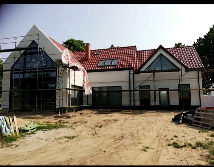 Nowoczesny dom jednorodzinny z czerwonym dachem w trakcie budowy, widoczne rusztowania i zabezpieczenia okien folią, teren wokół domu niezagospodarowany.