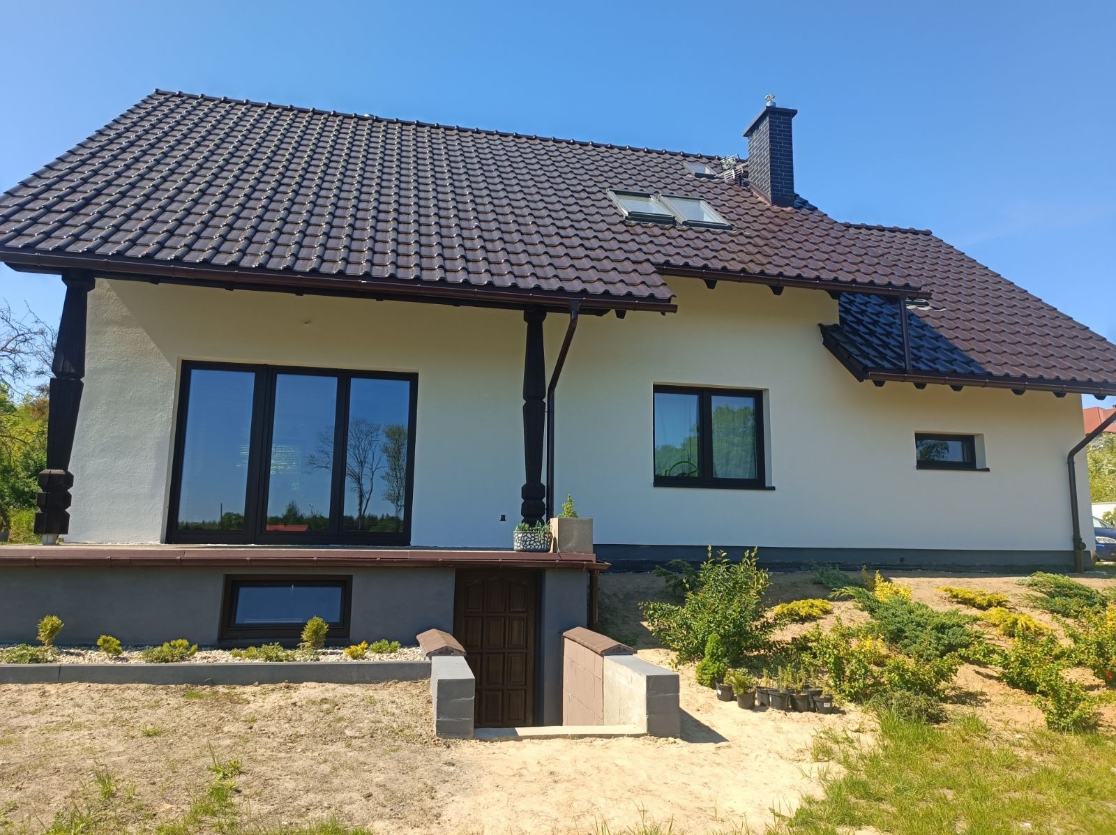 Biały dom z brązowym dachem, ciemnymi oknami i wejściem do piwnicy z betonowymi ściankami, otoczony zielenią i piaszczystym terenem.