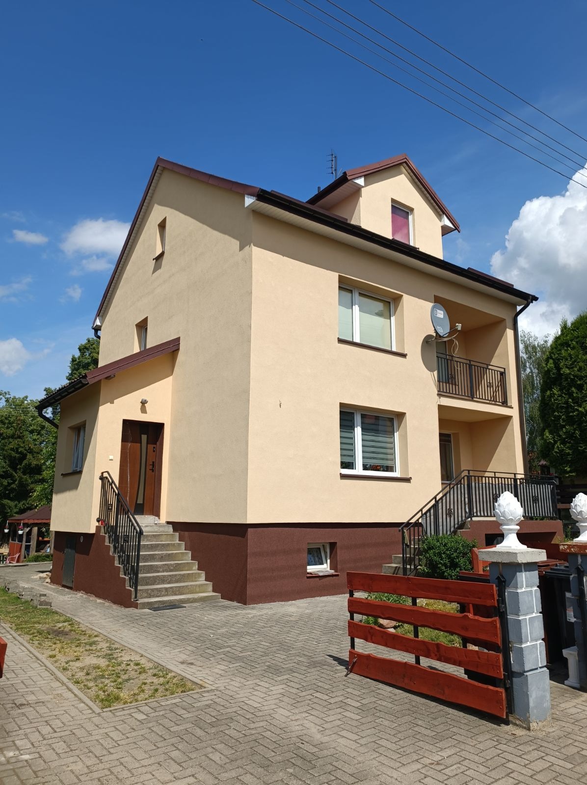 Dwukondygnacyjny dom z beżową elewacją, brązowym cokołem i balkonem, widoczny z poziomu gruntu w słoneczny dzień.