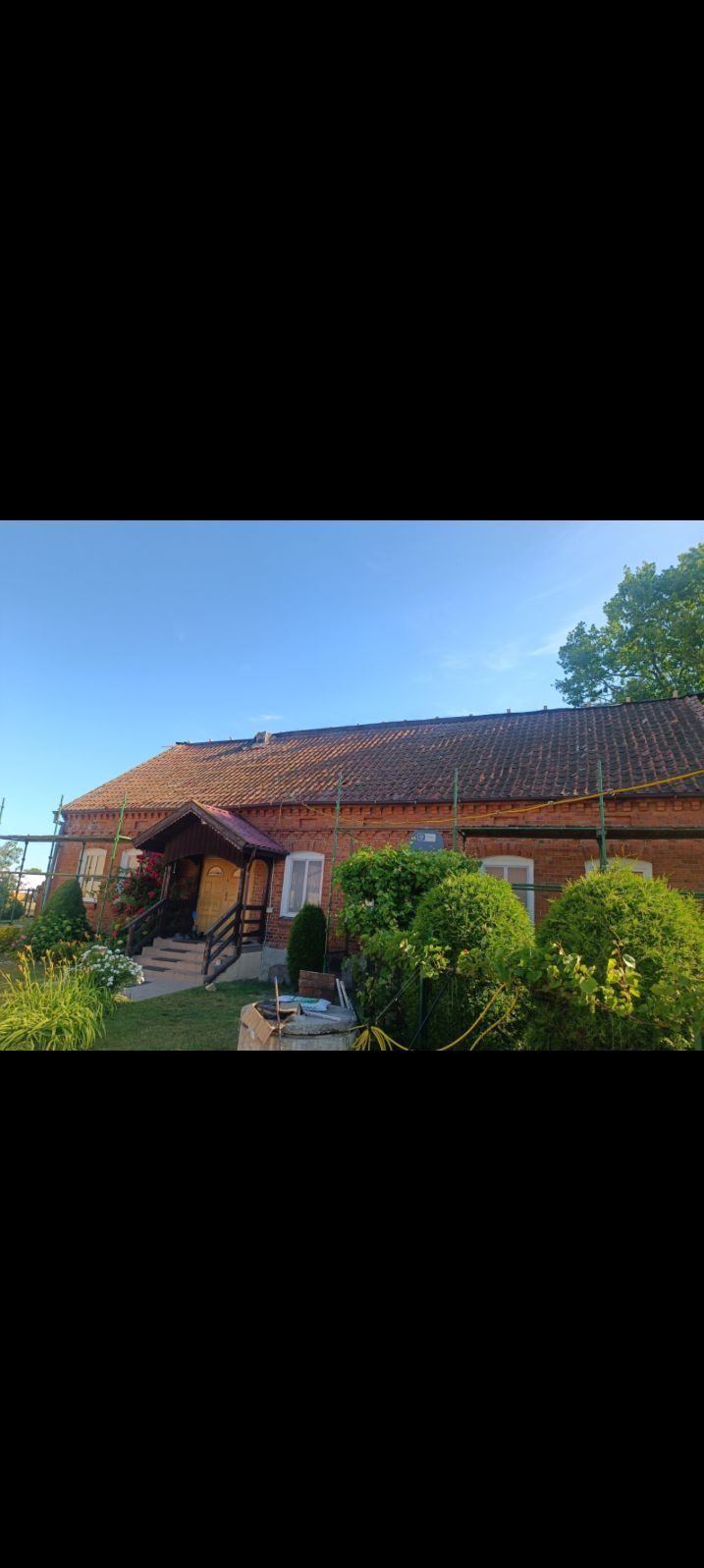 Ceglany dom z czerwonym dachem i drewnianym gankiem w trakcie renowacji, widoczne rusztowania i bujna roślinność w ogrodzie.