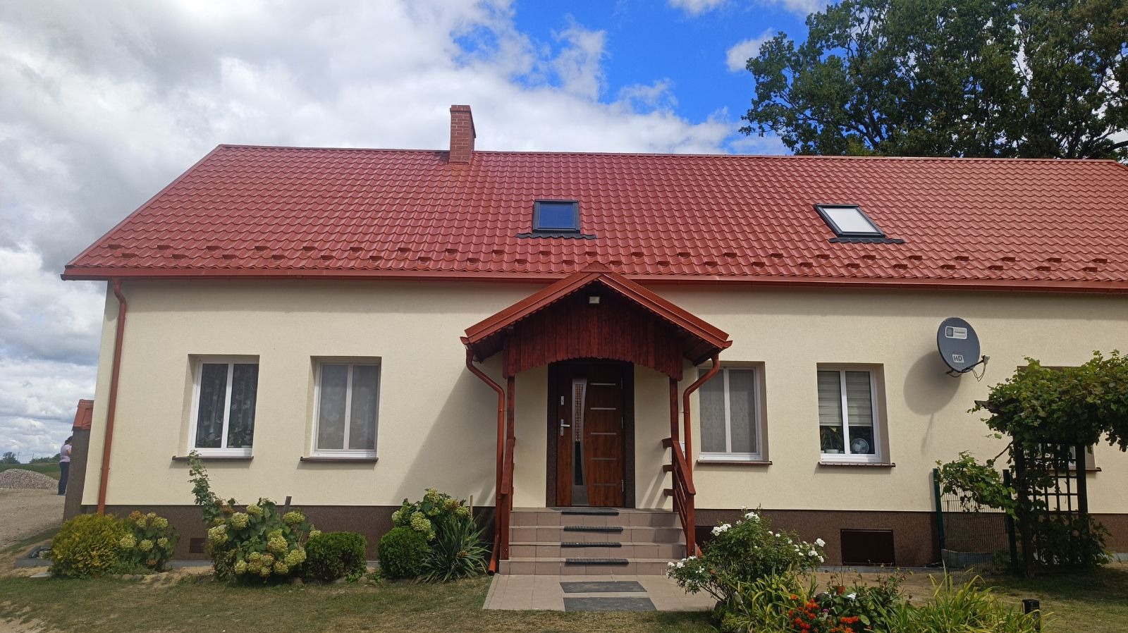 Wykończony dom jednorodzinny z czerwoną dachówką, kremową elewacją, oknami dachowymi i zadaszonym wejściem z drewnianymi elementami, otoczony zielenią w słoneczny dzień.