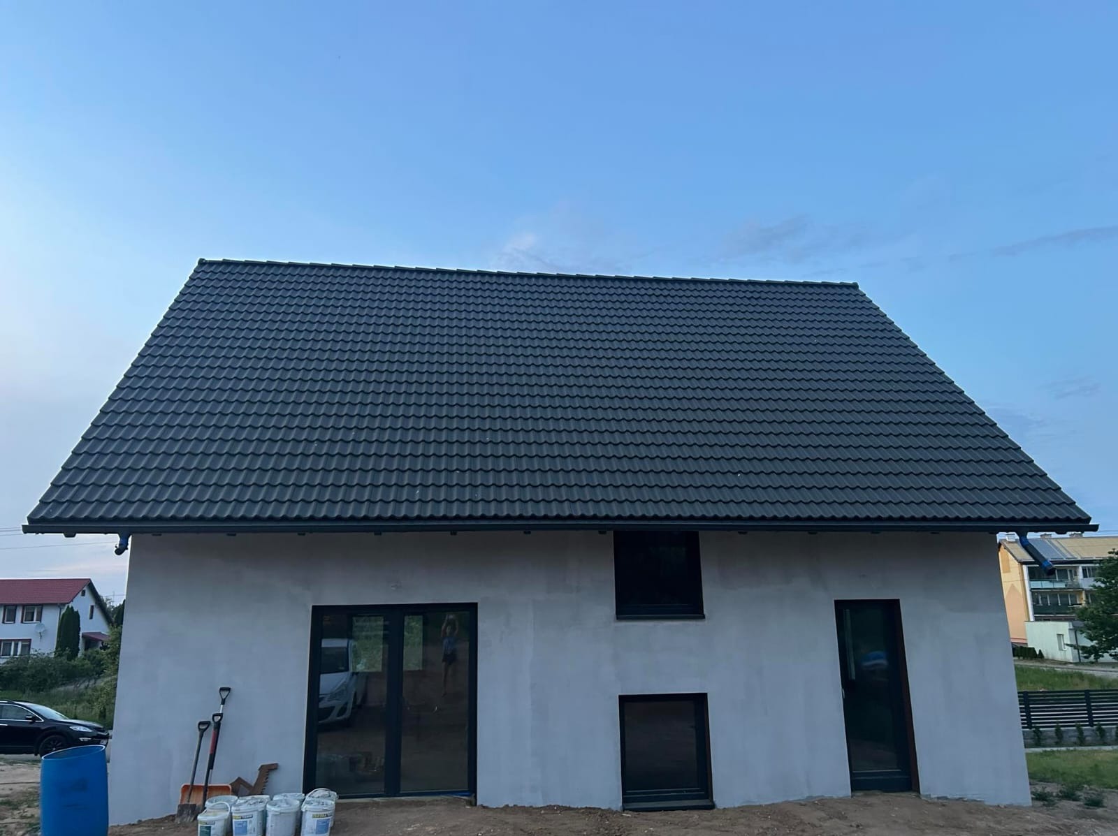 Nowoczesny dom jednorodzinny w trakcie budowy, z ciemną dachówką i surową elewacją, widoczne narzędzia i puste wiadra na ziemi.