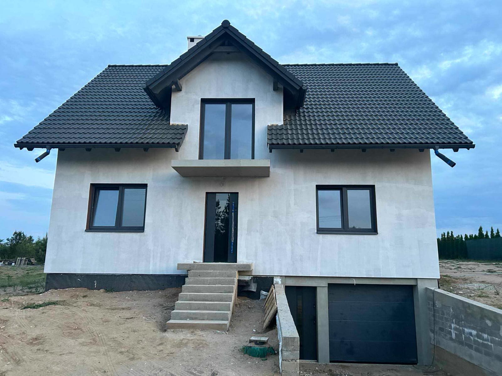 Surowy stan domu murowanego z garażem w Giżycku, z ciemną dachówką i stolarką okienną, widoczne schody wejściowe i teren w trakcie zagospodarowania.