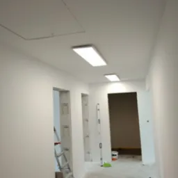 Korytarz w trakcie malowania na biało, widoczna drabina, poziomica, narzędzia malarskie i sufit z oświetleniem LED.