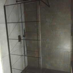 Lukasz Stadnik - Nowoczesna kabina prysznicowa z czarnymi profilami i deszczownicą, wykończona płytkami w odcieniach szarości i rdzy, oświetlona punktowo.