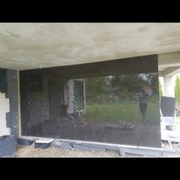 Lukasz Stadnik - Montaż ciemnych, błyszczących płytek z marmurkowym wzorem na elewacji budynku; widoczne niebieskie kliny dystansowe, styropianowe ocieplenie i odbicie osoby w płytkach.