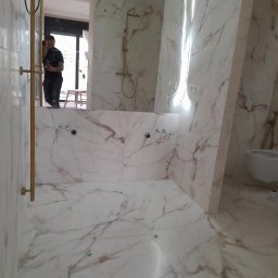 Lukasz Stadnik - Wykończona łazienka z marmurowymi płytkami w odcieniach bieli i beżu, złota armatura i oświetlenie LED, widoczne przygotowane otwory na baterie i syfon, odbicie pracownika w lustrze.