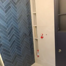 Ściana łazienki wyłożona niebieską glazurą w jodełkę, z wnęką na kosmetyki i widocznymi przyłączami wodnymi, oraz fragmentem szafki w kolorze granatowym.