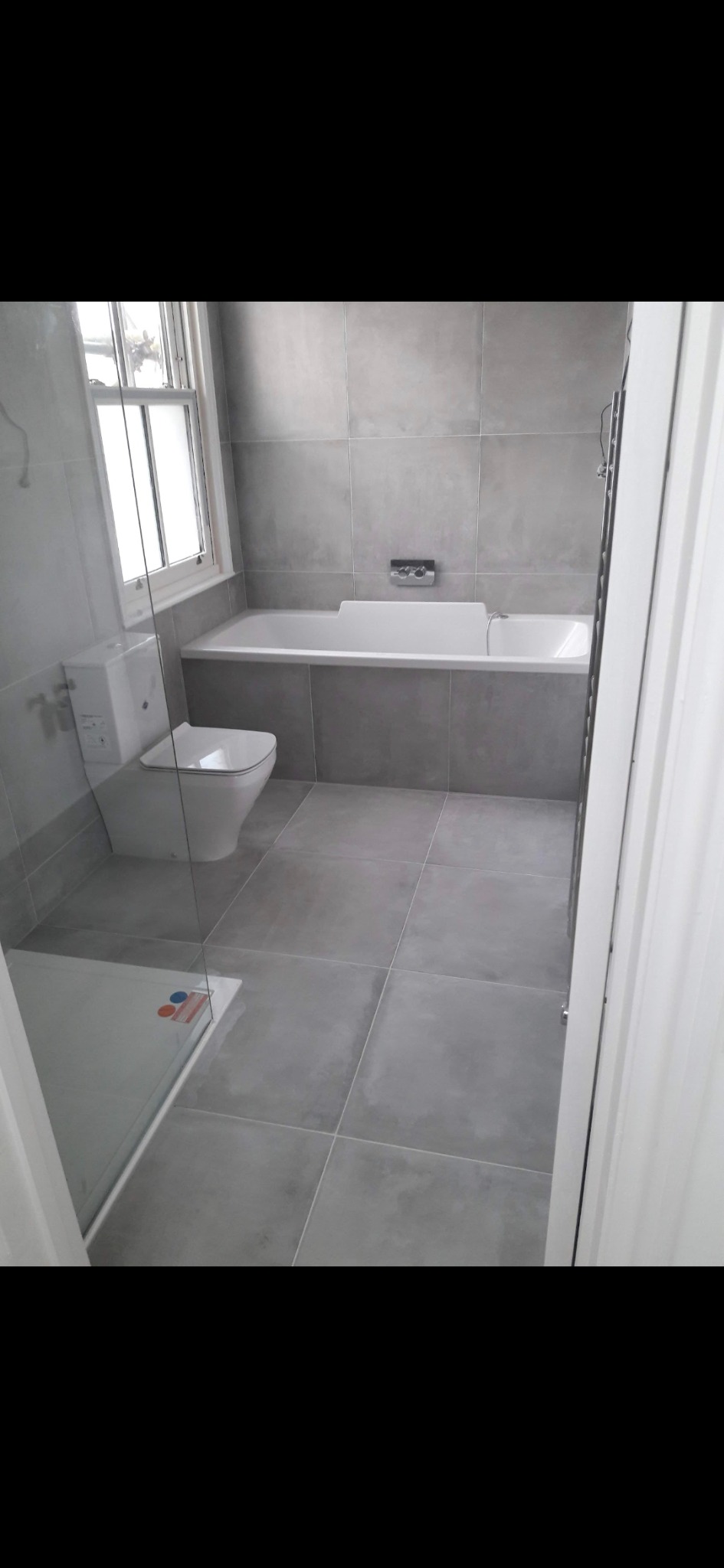 Nowoczesna łazienka wykończona w szarościach: płytki ceramiczne na ścianach i podłodze, biała wanna zabudowana, toaleta typu compact i kabina prysznicowa z transparentną szybą.