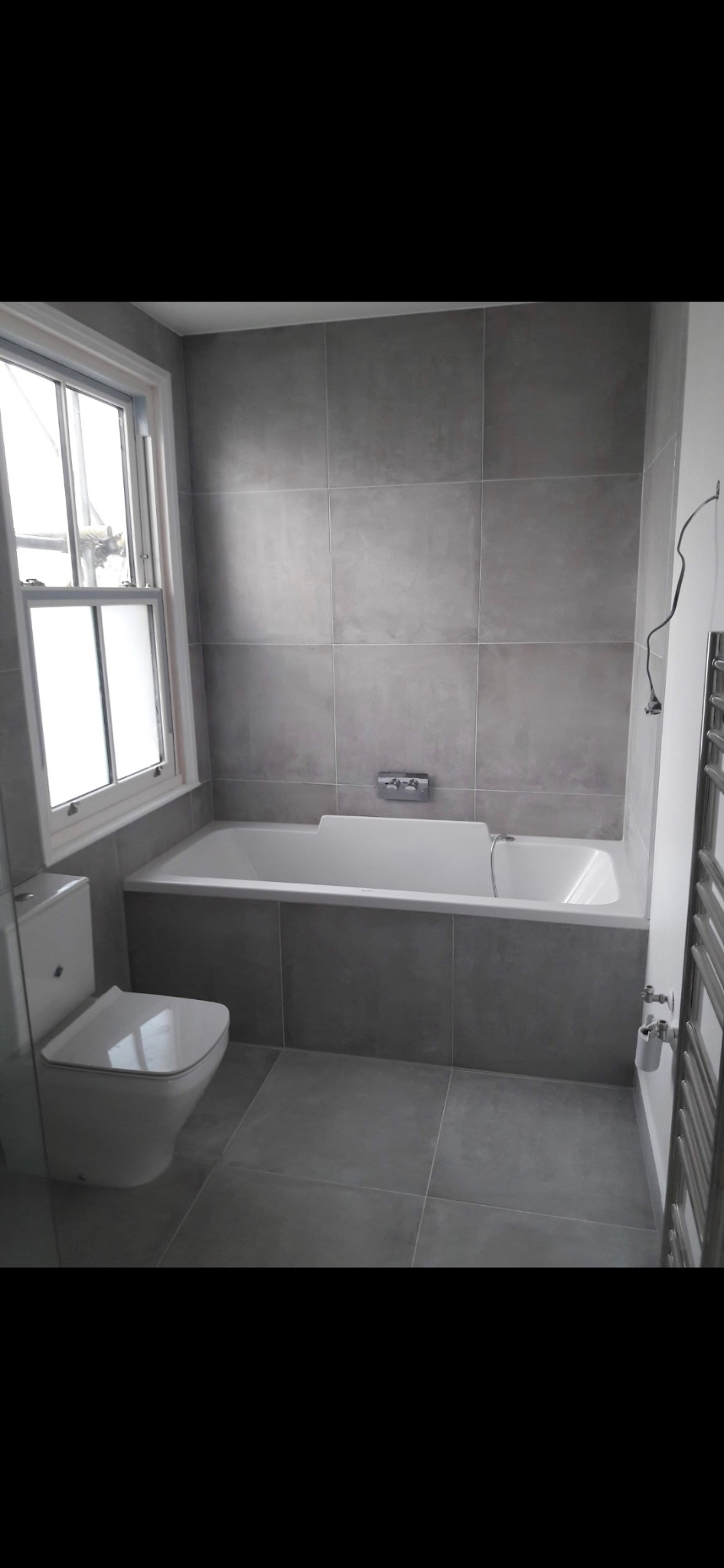 Wykończona łazienka w odcieniach szarości z białą wanną zabudowaną płytkami i toaletą typu compact, widoczna przez szybę kabiny prysznicowej, z oknem w tle.