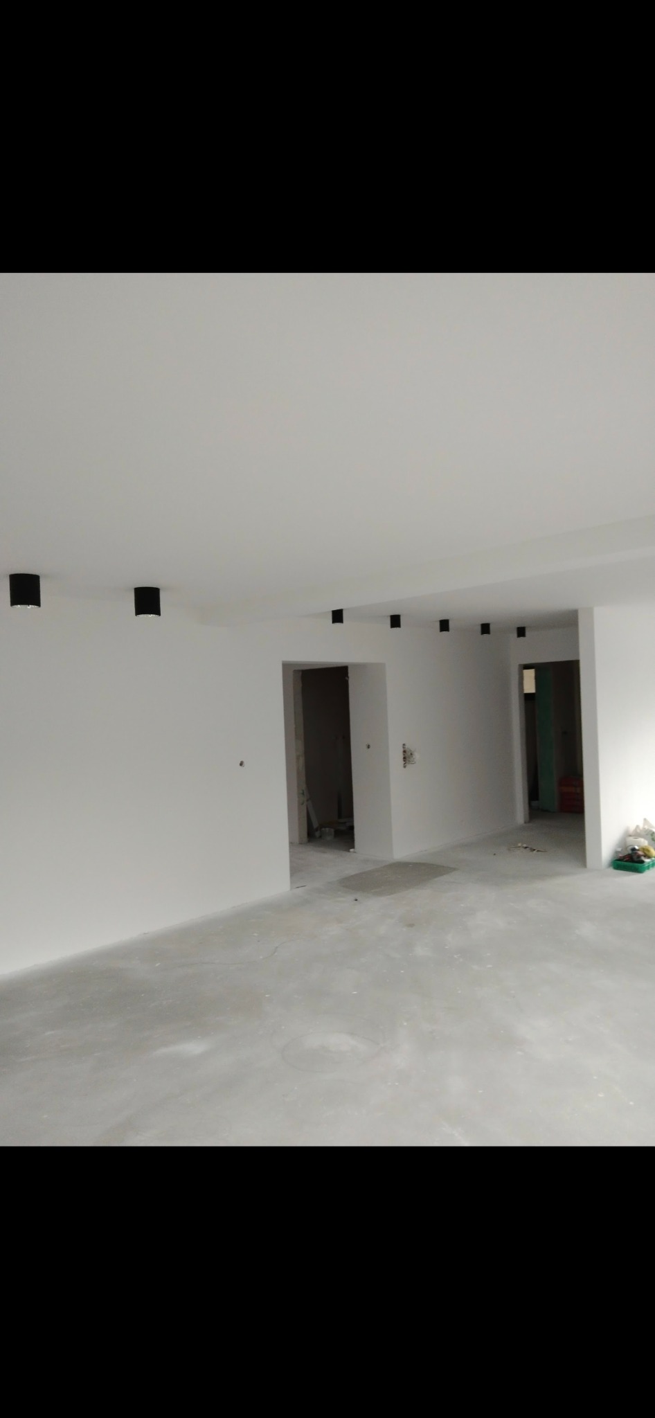 Wnętrze w trakcie remontu: świeżo pomalowane na biało ściany i sufit, widoczne czarne, minimalistyczne lampy sufitowe i surowa betonowa podłoga przed wykończeniem.