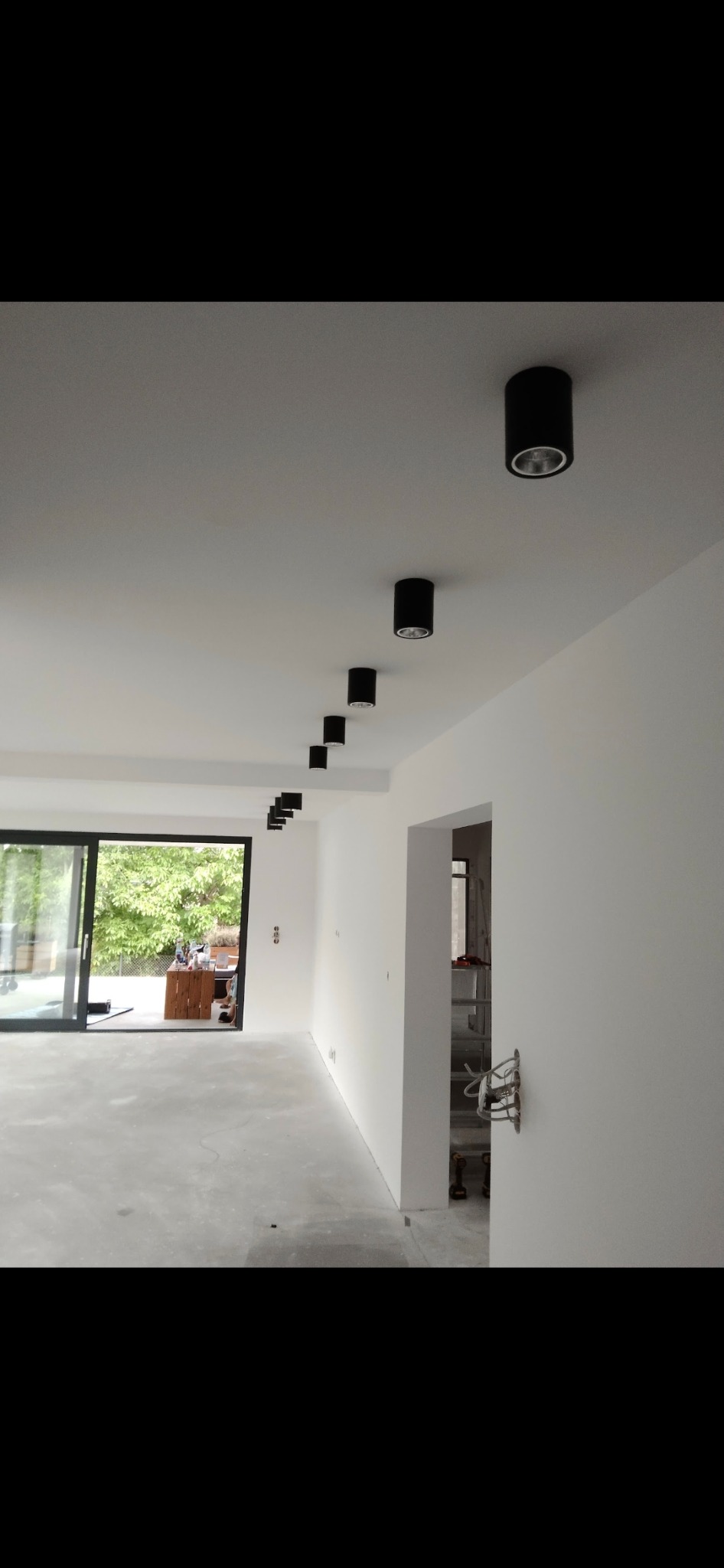 Nowoczesne oświetlenie sufitowe w rzędzie, czarne tuby, białe ściany i sufit, surowy beton na podłodze, duże przesuwne okno z widokiem na zieleń, wnętrze w trakcie wykańczania.