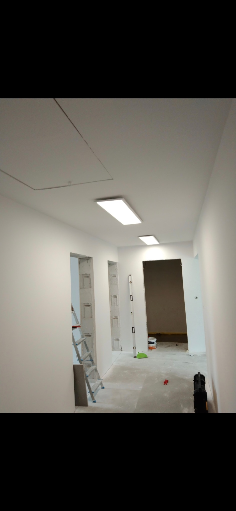 Korytarz w trakcie malowania na biało, widoczna drabina, poziomica, narzędzia malarskie i sufit z oświetleniem LED.