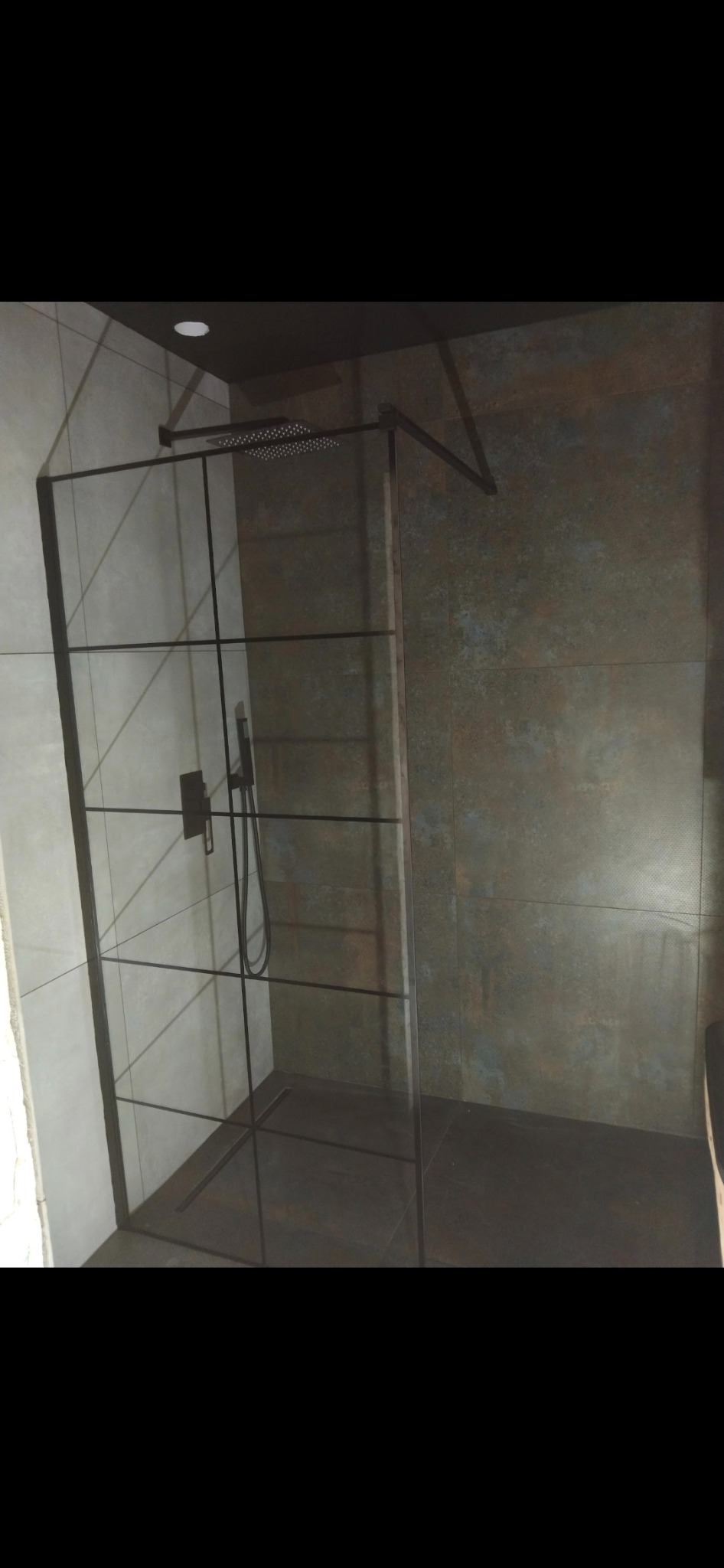 Nowoczesna kabina prysznicowa z czarnymi profilami i deszczownicą, wykończona płytkami w odcieniach szarości i rdzy, oświetlona punktowo.