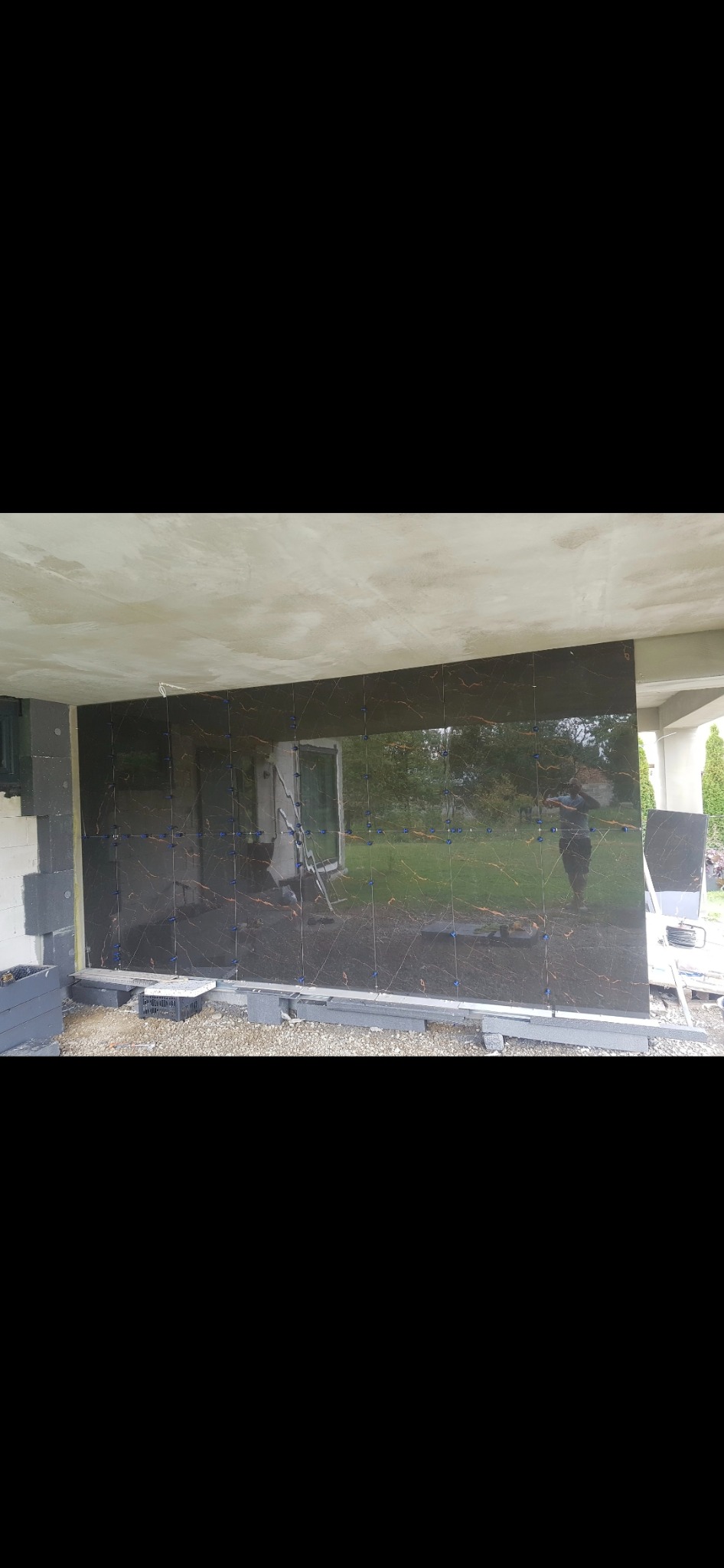 Montaż ciemnych, błyszczących płytek z marmurkowym wzorem na elewacji budynku; widoczne niebieskie kliny dystansowe, styropianowe ocieplenie i odbicie osoby w płytkach.