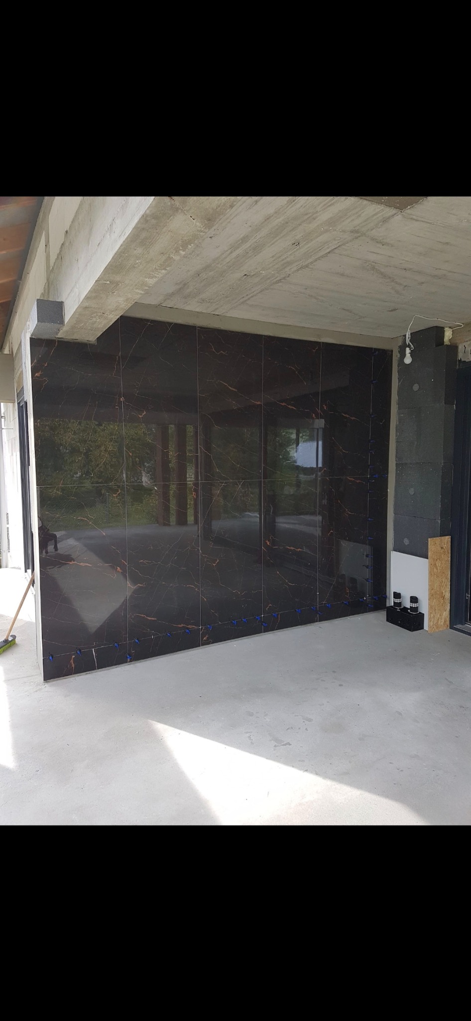 Ściana wyłożona ciemnymi płytkami imitującymi marmur z widocznymi niebieskimi krzyżykami dystansowymi, surowy betonowy sufit, stan surowy budynku.