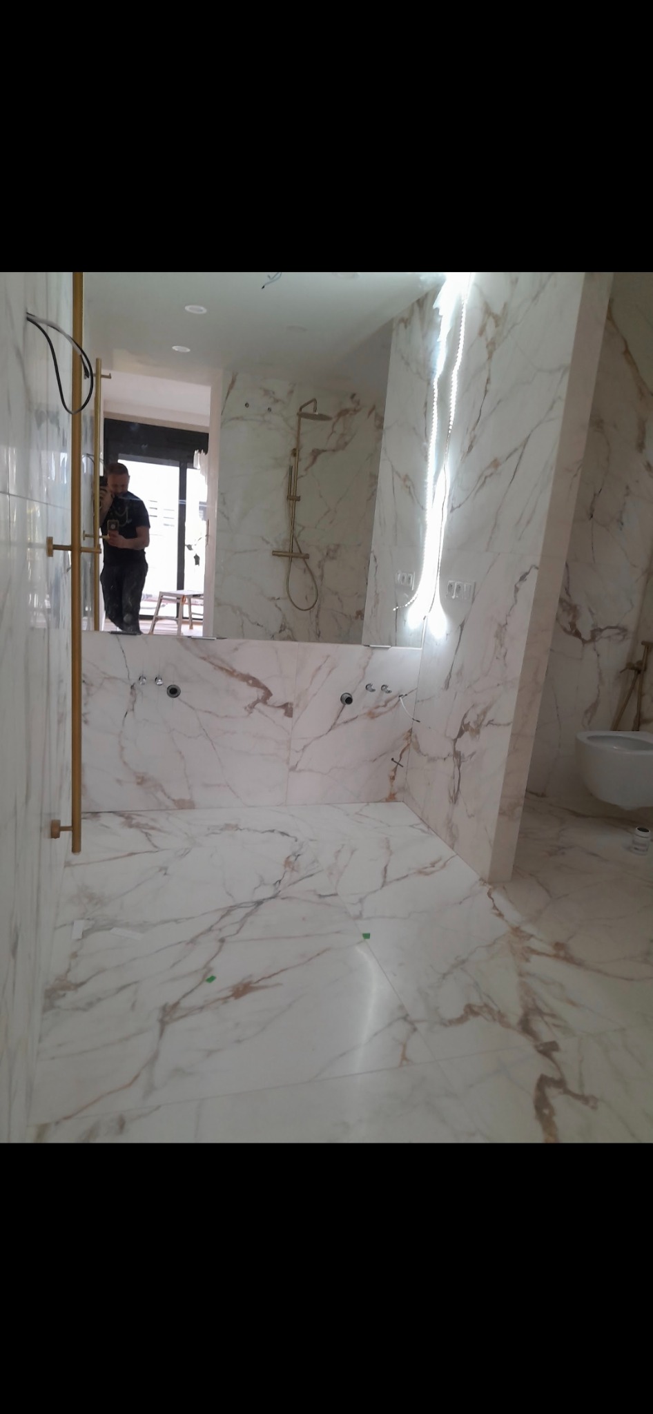 Wykończona łazienka z marmurowymi płytkami w odcieniach bieli i beżu, złota armatura i oświetlenie LED, widoczne przygotowane otwory na baterie i syfon, odbicie pracownika w lustrze.