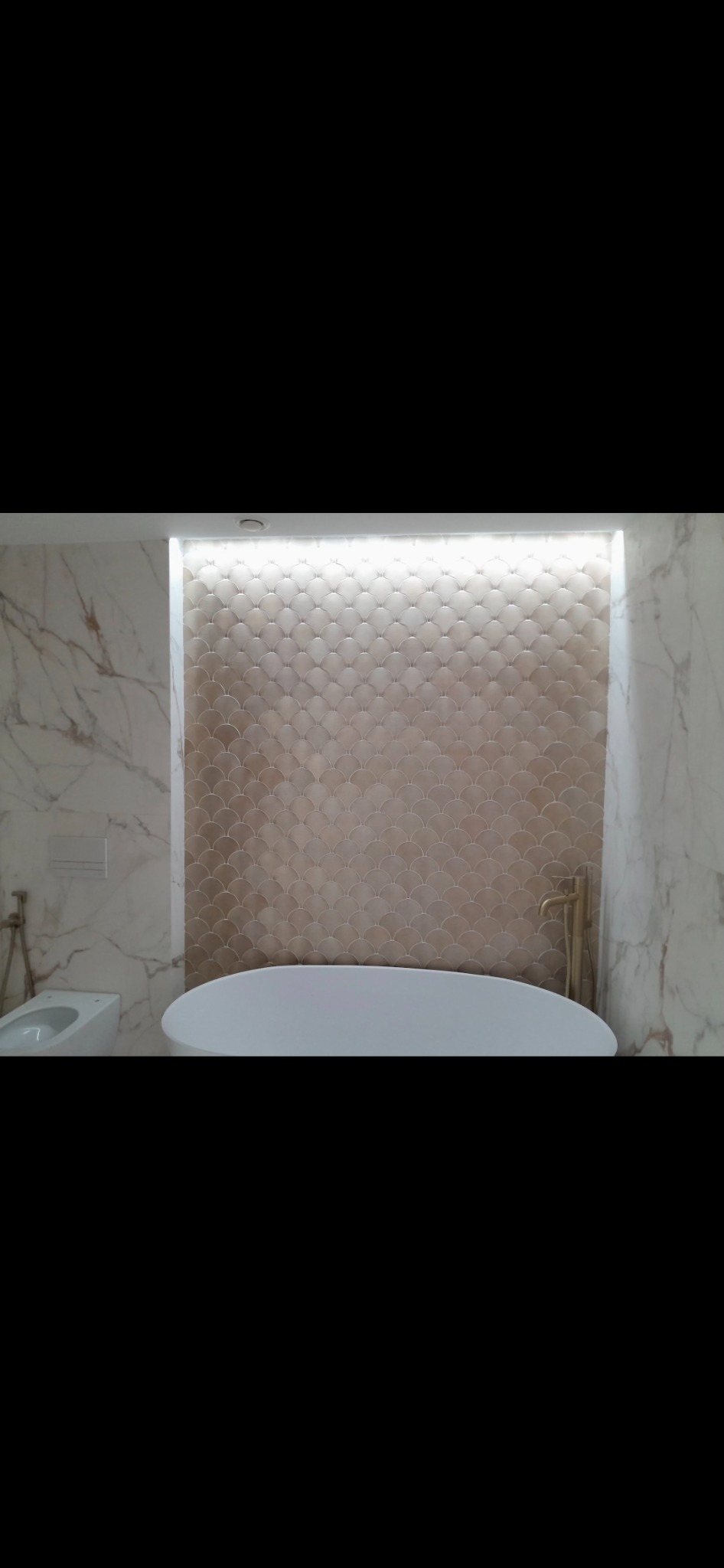 Nowoczesna łazienka z wolnostojącą wanną, złotą armaturą i ścianą wyłożoną beżowymi płytkami w kształcie rybiej łuski, oświetlona listwą LED.