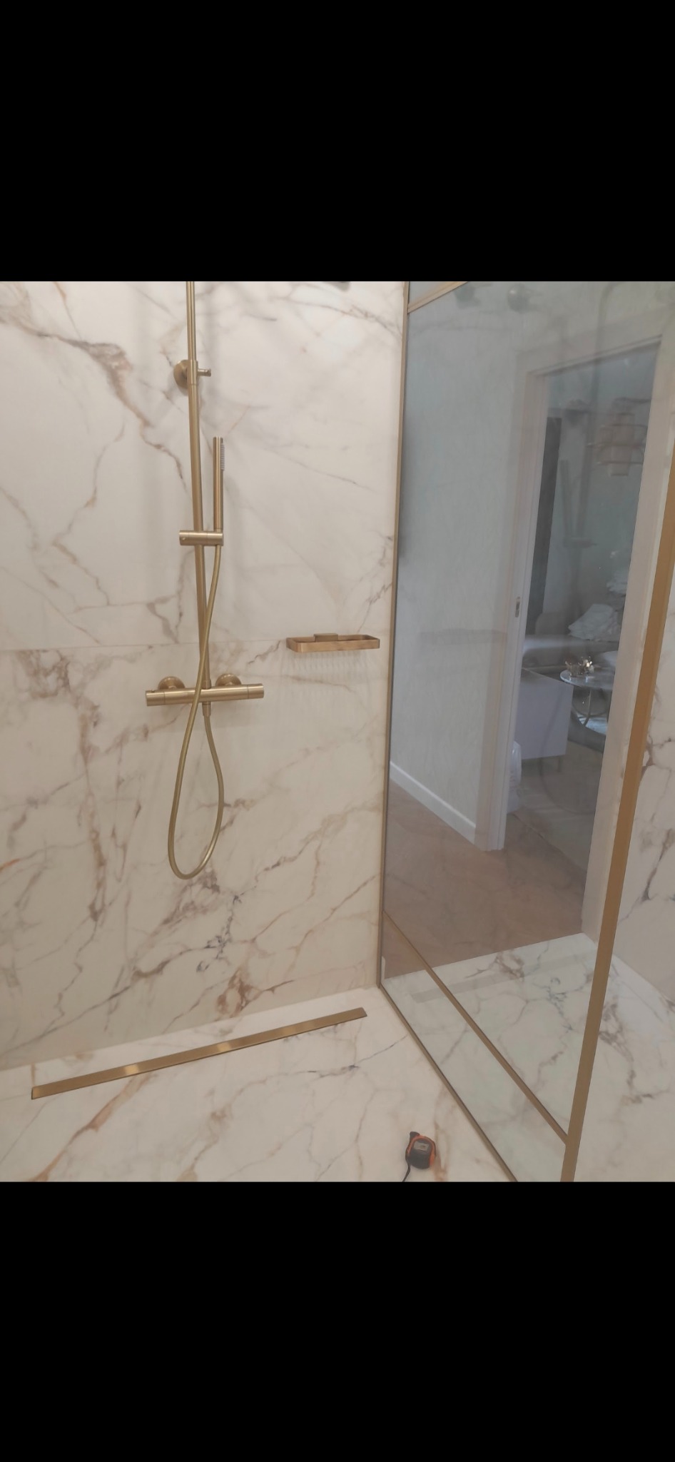 Kabina prysznicowa z marmurowymi płytkami w odcieniach bieli i beżu, złota armatura i listwy wykończeniowe, widoczny odpływ liniowy i częściowo widoczne pomieszczenie za szklaną ścianką.