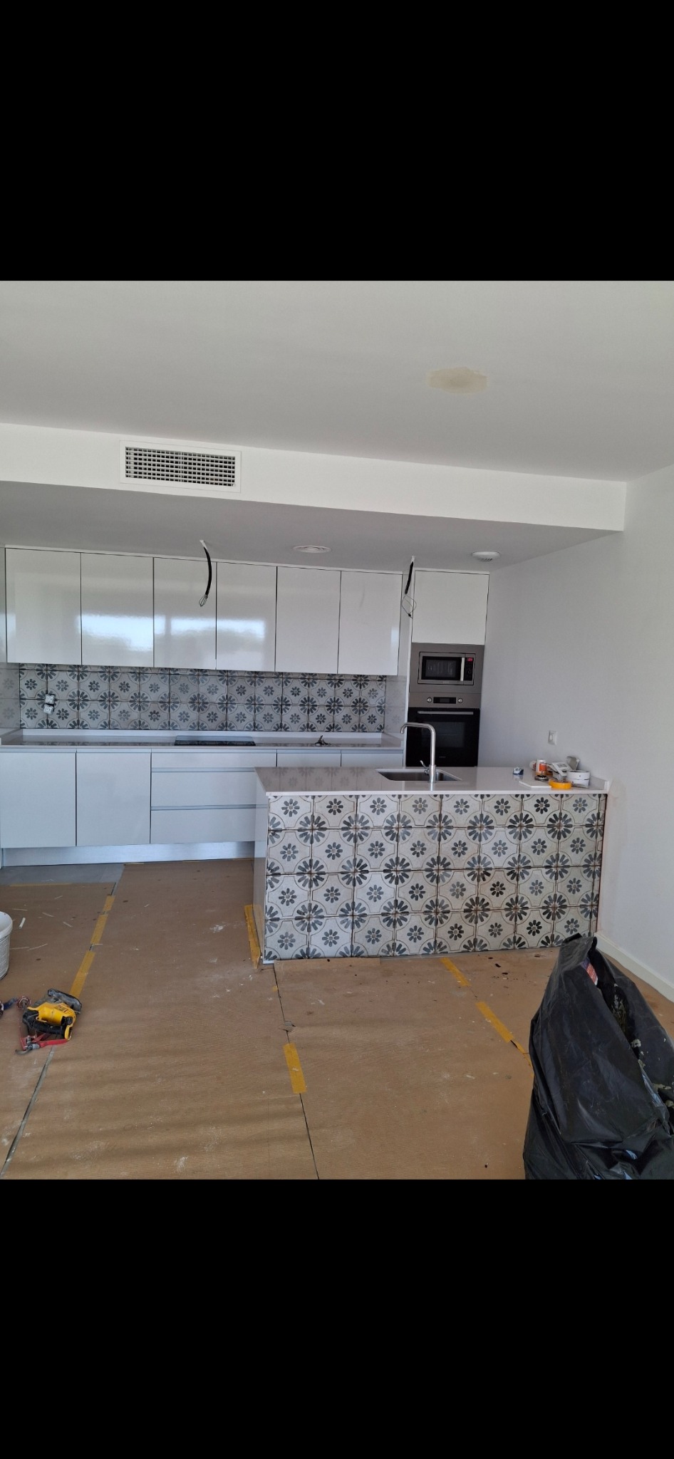 Wnętrze kuchni w trakcie wykańczania: białe szafki z wzorzystymi płytkami nad blatem, wyspa kuchenna z identycznymi płytkami, zabezpieczona podłoga, narzędzia i worek na śmieci w tle.