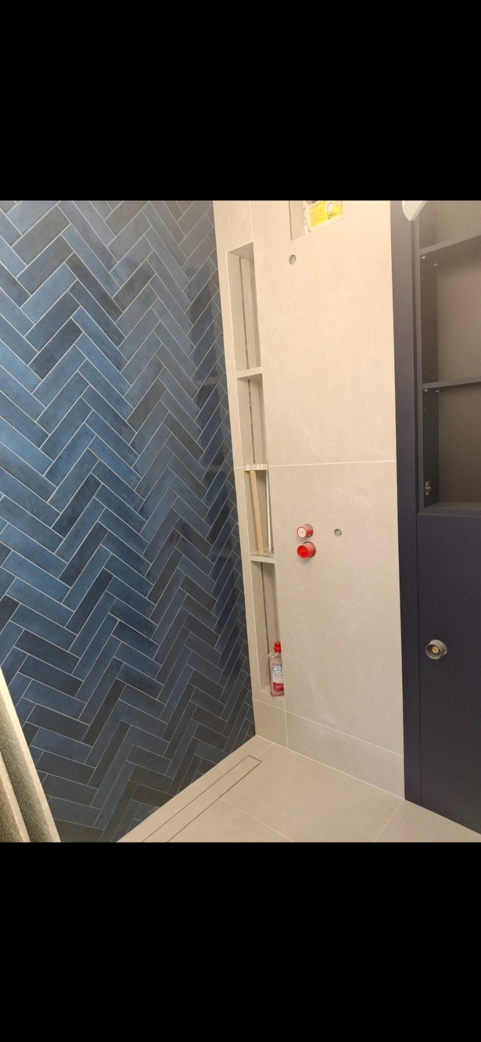 Ściana łazienki wyłożona niebieską glazurą w jodełkę, z wnęką na kosmetyki i widocznymi przyłączami wodnymi, oraz fragmentem szafki w kolorze granatowym.