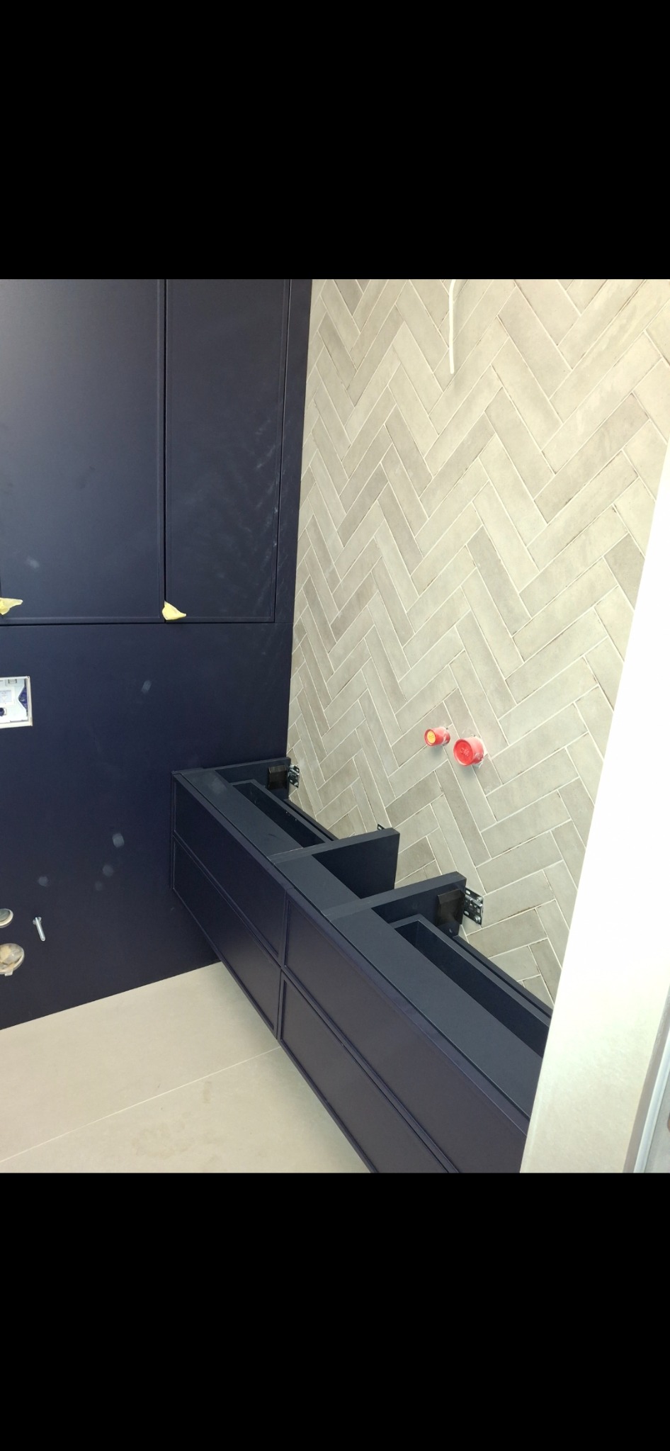 Niebieska szafka łazienkowa z szufladami, zamontowana na ścianie, z widocznymi elementami instalacji wodnej, na tle ściany wyłożonej jasnymi płytkami w jodełkę.