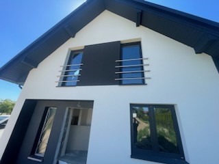 Nowoczesny dom z białą elewacją, czarnymi ramami okiennymi i grafitowym panelem dekoracyjnym między oknami na piętrze, z metalowymi balustradami.