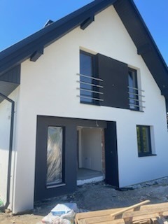 Nowoczesny dom jednorodzinny z białą elewacją i czarnymi detalami wykończeniowymi, widoczne okna z metalowymi barierkami balkonowymi, teren wokół domu w trakcie prac wykończeniowych.