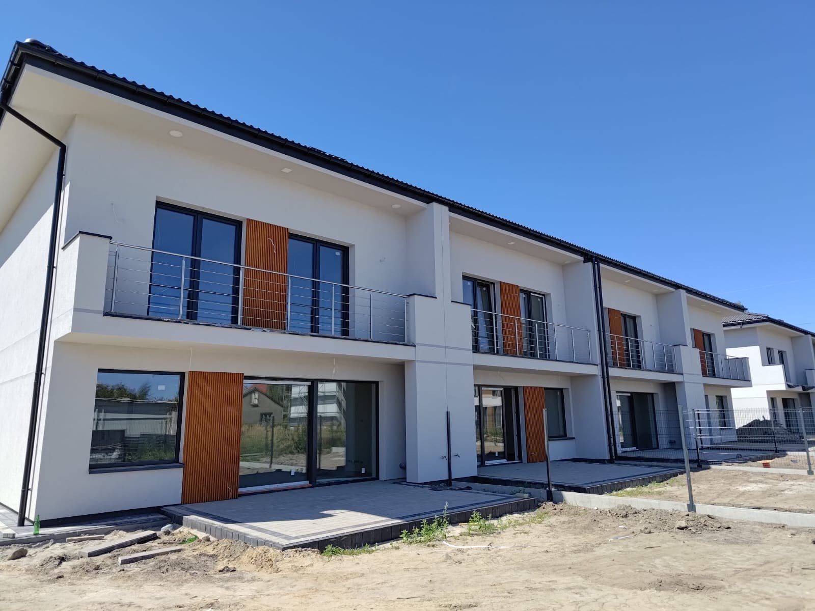 Nowoczesne domy w zabudowie szeregowej z białymi elewacjami, drewnianymi panelami dekoracyjnymi i balkonami ze stali nierdzewnej, w trakcie prac wykończeniowych.