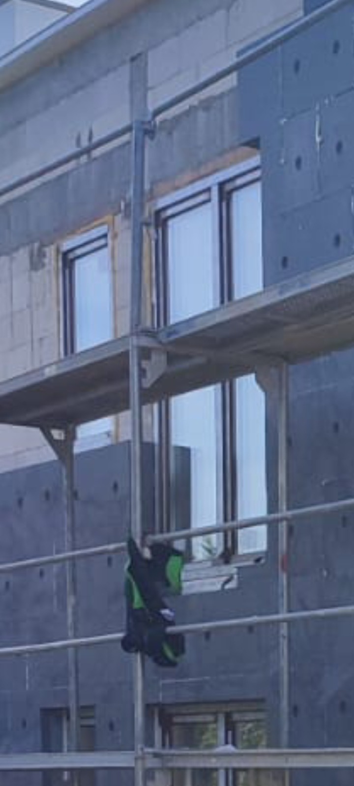 Budynek w trakcie ocieplania, widoczne rusztowanie, szare płyty izolacyjne z widocznymi otworami montażowymi oraz fragment ściany z bloczków, okna z ciemnymi ramami.