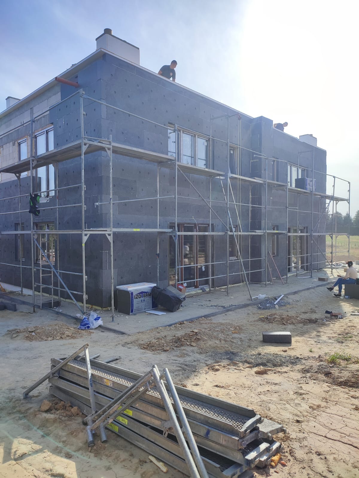 Dwupiętrowy dom w trakcie ocieplania, pokryty szarymi płytami izolacyjnymi, otoczony rusztowaniami, z pracownikami na dachu i przy elewacji, widoczne materiały izolacyjne IZO-TERM oraz składowane...
