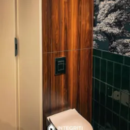 Nowoczesna toaleta z drewnianą obudową stelaża, zielonymi płytkami i tapetą w motywy drzew, minimalistyczny przycisk spłukujący.