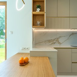 iNTEGRITI - Nowoczesna kuchnia z drewnianym blatem, marmurowym backsplash'em i otwartymi półkami z dekoracjami. Widok na ogród przez okno. Oświetlenie LED podkreśla detale.