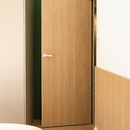 iNTEGRITI - Otwarta, fornirowana dębem drzwi wewnętrzne z białą klamką, na tle zielonej ściany i beżowej szafy w minimalistycznym wnętrzu. Widoczny fragment łóżka.