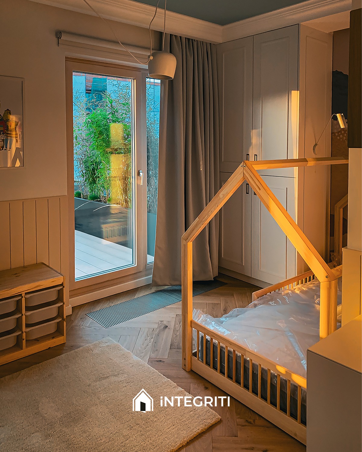 Pokój dziecięcy z drewnianym łóżkiem w kształcie domku, jasnym dywanem i szafką z szufladami, parkiet ułożony w jodełkę, widok na taras przez przeszklone drzwi balkonowe.