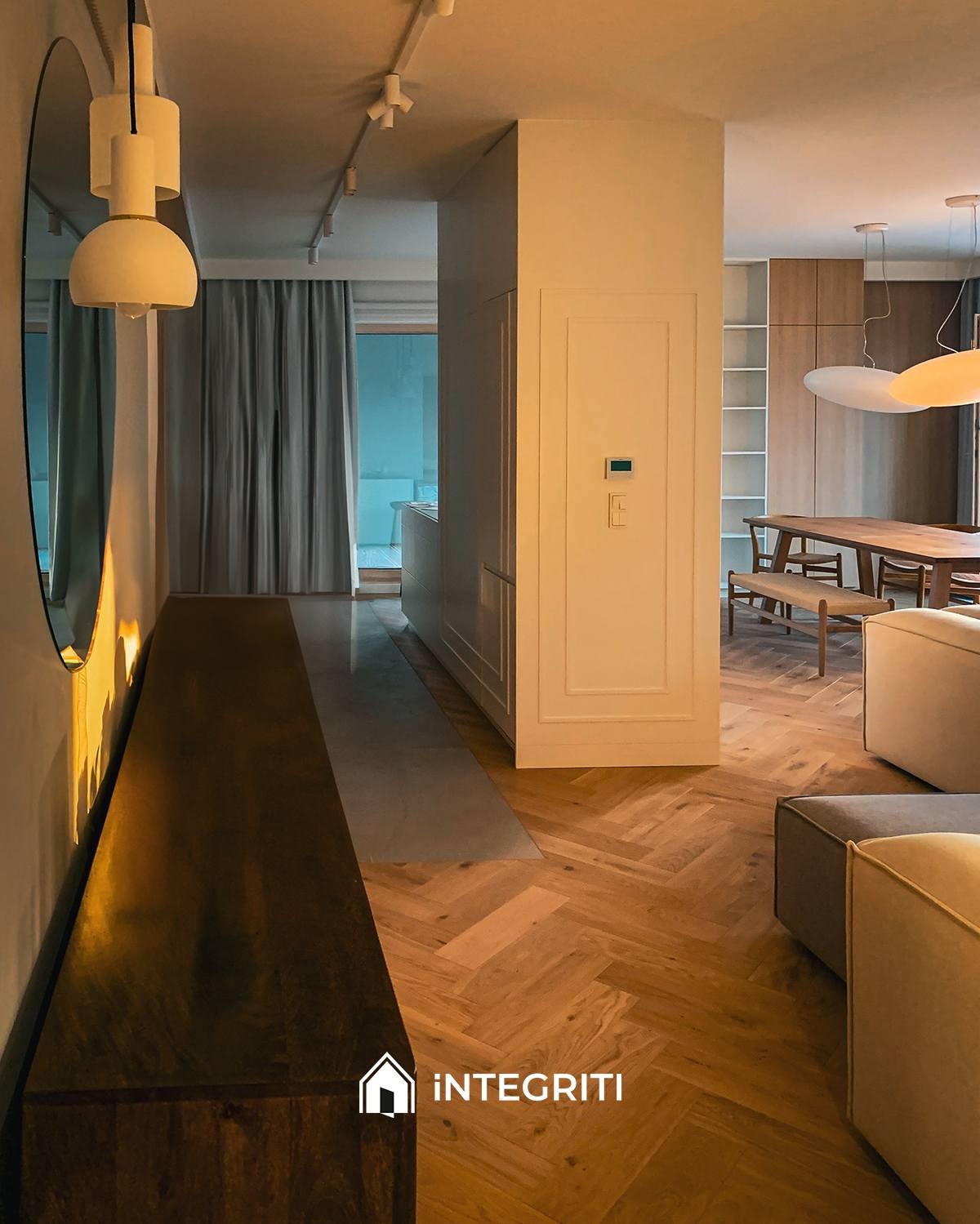 Nowoczesne wnętrze z parkietem ułożonym w jodełkę, ciemnym drewnianym blatem wzdłuż ściany, okrągłym lustrem i lampami sufitowymi. W tle widoczna jadalnia z drewnianym stołem i krzesłami.