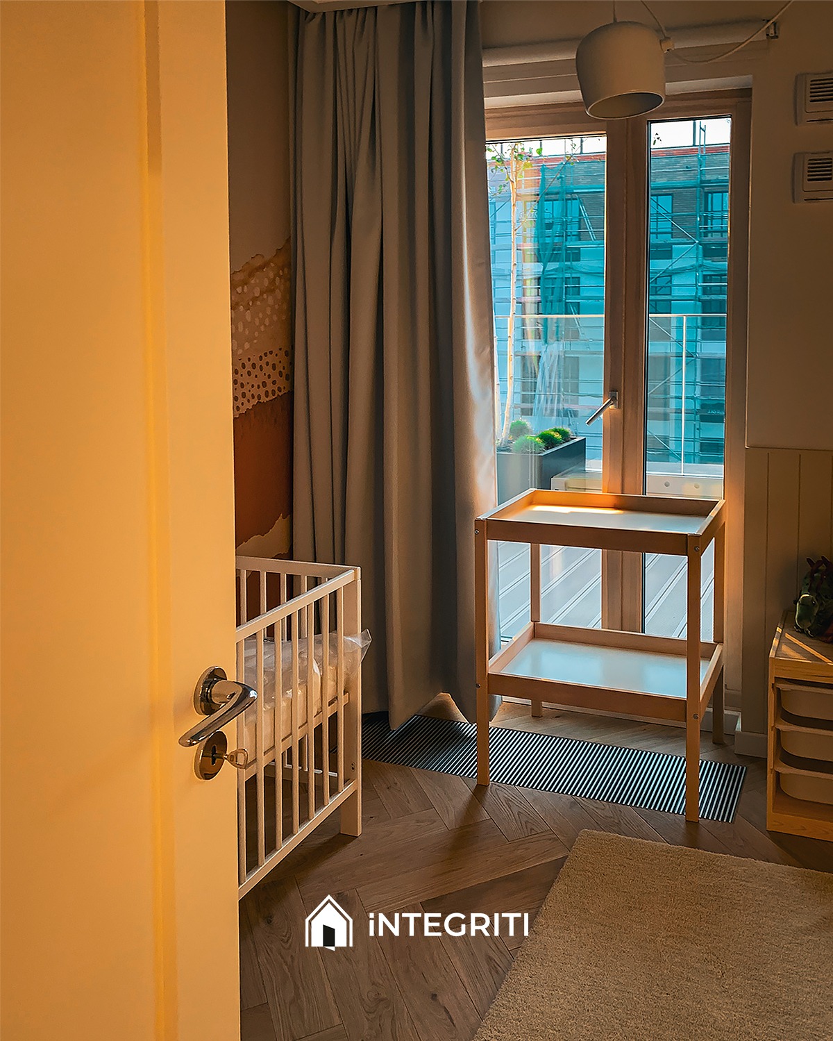 Umeblowany pokój dziecięcy z łóżeczkiem, przewijakiem i zasłoniętym oknem, widok zza uchylonych drzwi.