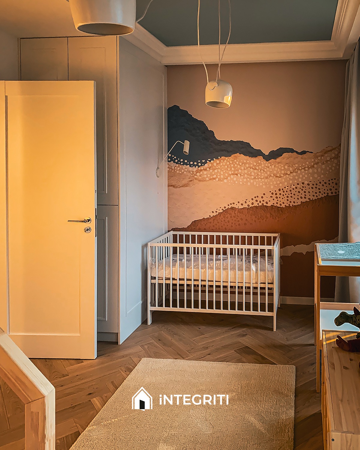 Pokój dziecięcy z białym łóżeczkiem, malowaną ścianą w piaskowe góry i beżowym dywanem z logo firmy na podłodze z parkietu ułożonego w jodełkę.