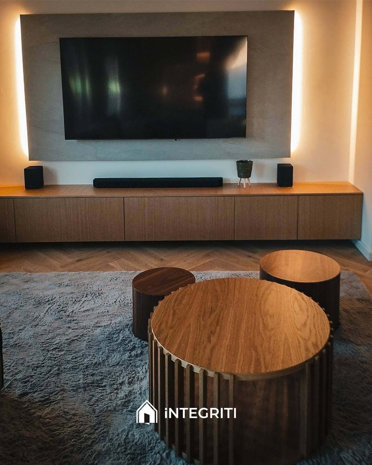 Salon z telewizorem na ścianie podświetlonej listwami LED, drewnianą szafką RTV, soundbarem i głośnikami, dywanem w odcieniach szarości oraz zestawem okrągłych stolików kawowych z drewna.