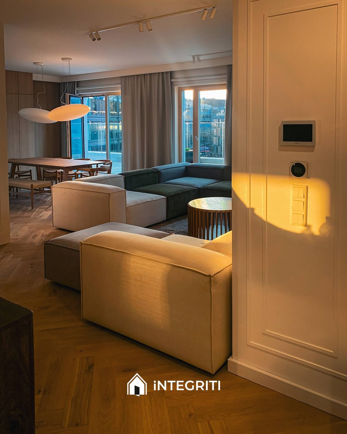 Jasny salon z parkietem ułożonym w jodełkę, modułowa sofa w odcieniach beżu i zieleni, okrągły stolik kawowy z drewnianą podstawą oraz widok na miasto zza okien.