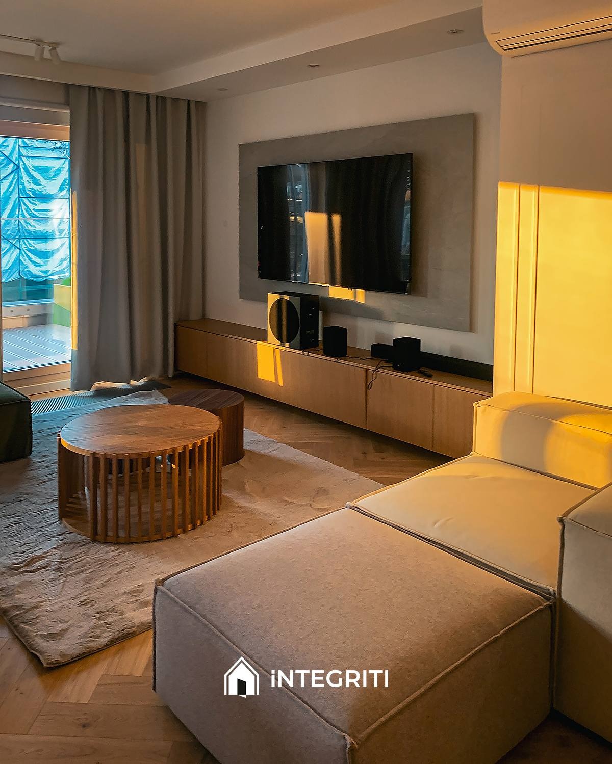 Nowoczesny salon z drewnianą podłogą i okrągłym stolikiem kawowym, minimalistyczna szafka RTV pod telewizorem, ciepłe światło zachodzącego słońca wpadające przez okno balkonowe, logo firmy na sofie.