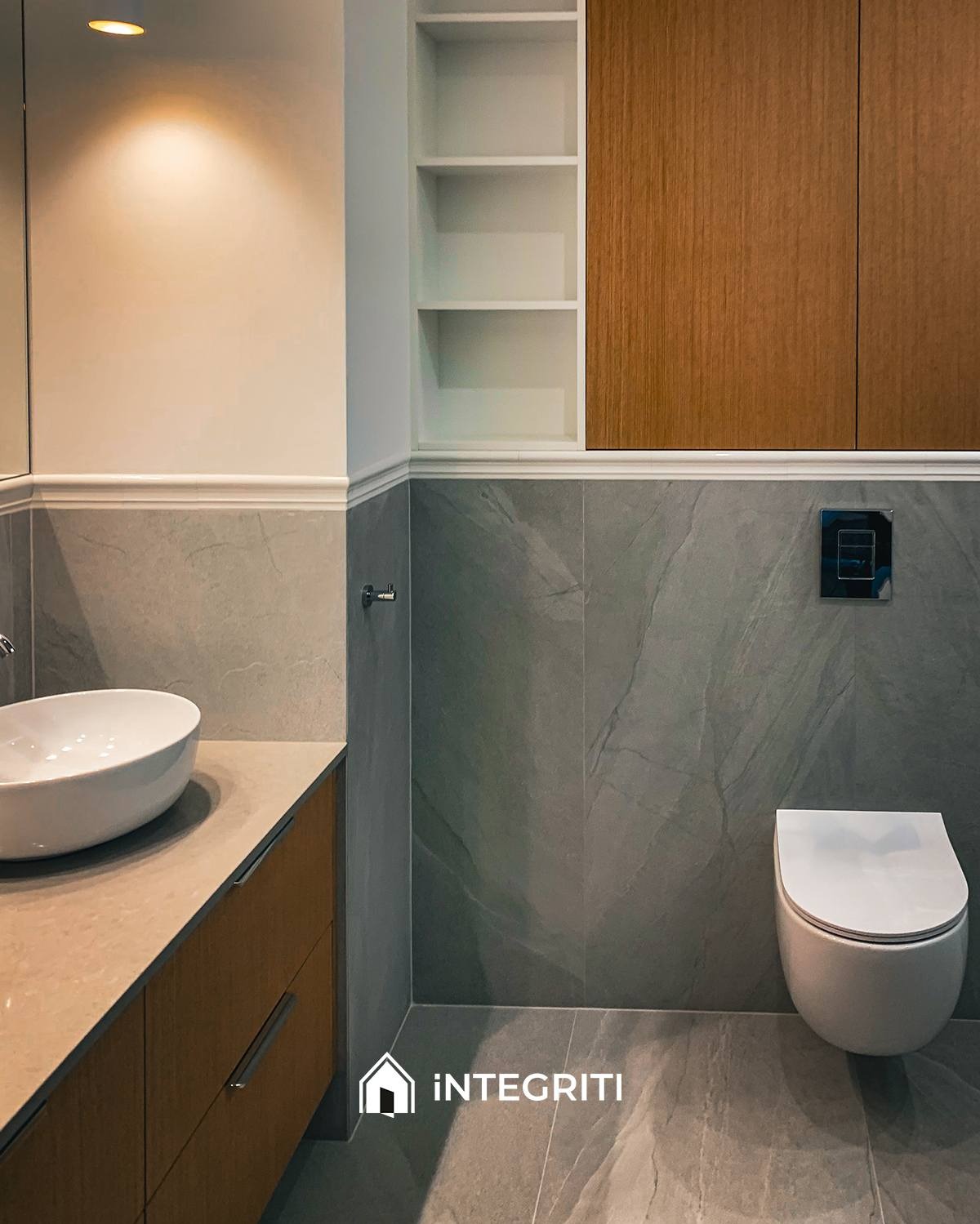 Nowoczesna łazienka z umywalką nablatową, toaletą wiszącą i szarą marmurową okładziną ścienną, oświetlona punktowym światłem, z zabudową meblową z drewnianymi frontami i białymi półkami.